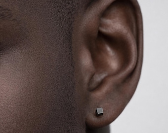 May include: A close-up of a person's ear with a small, square, dark gray stud earring. The earring is positioned on the earlobe. The skin tone is dark.