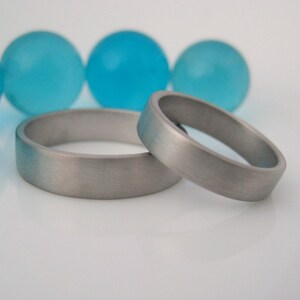 May include: Two silver wedding bands, one slightly larger than the other, are lying on a white surface. The bands have a brushed finish and are positioned side by side.