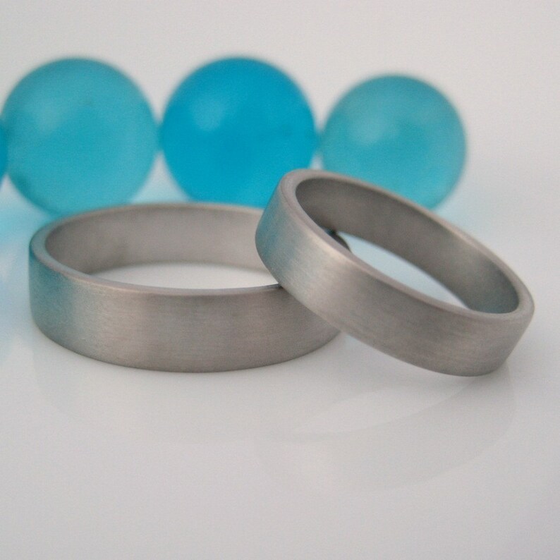 May include: Two silver wedding bands, one slightly larger than the other, are lying on a white surface. The bands have a brushed finish and are positioned side by side.