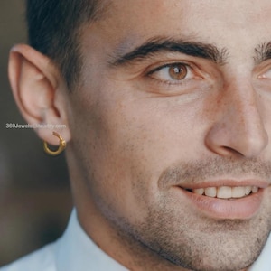 May include: Close-up of a person wearing a small, gold hoop earring. The earring is circular with a small decorative element. The person is smiling, and the earring is in focus. The website address 360JewelsElite.etsy.com is visible.