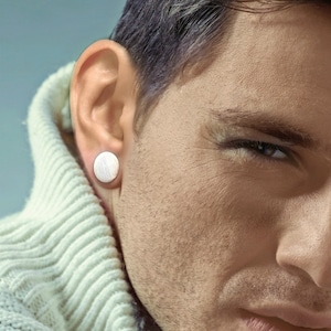 May include: A close-up of a man's face with a silver round earring in his left ear. The man is wearing a white sweater.