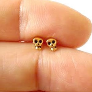 Op de afbeelding: Een paar kleine gouden schedel oorbel studs. De schedels zijn zwart en hebben een gedetailleerd ontwerp.