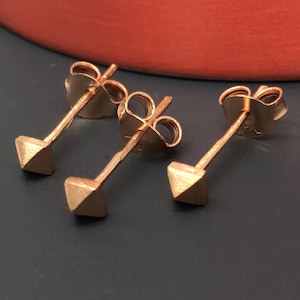 May include: Three gold-toned stud earrings with a triangular shape. The earrings are on a dark surface.