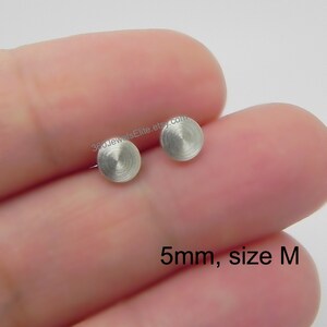 Puede incluir: Un par de pendientes de plata, cada uno con un diseño circular de metal cepillado. Los pendientes tienen un tamaño de 5 mm y están etiquetados como "talla M".