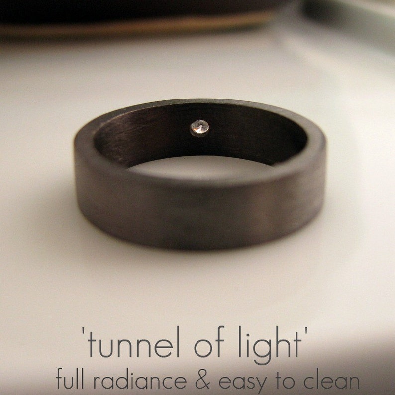 May include: A black ring with a single, small, clear gemstone set in the center. The ring is made of a smooth, dark material and has a simple, modern design.
