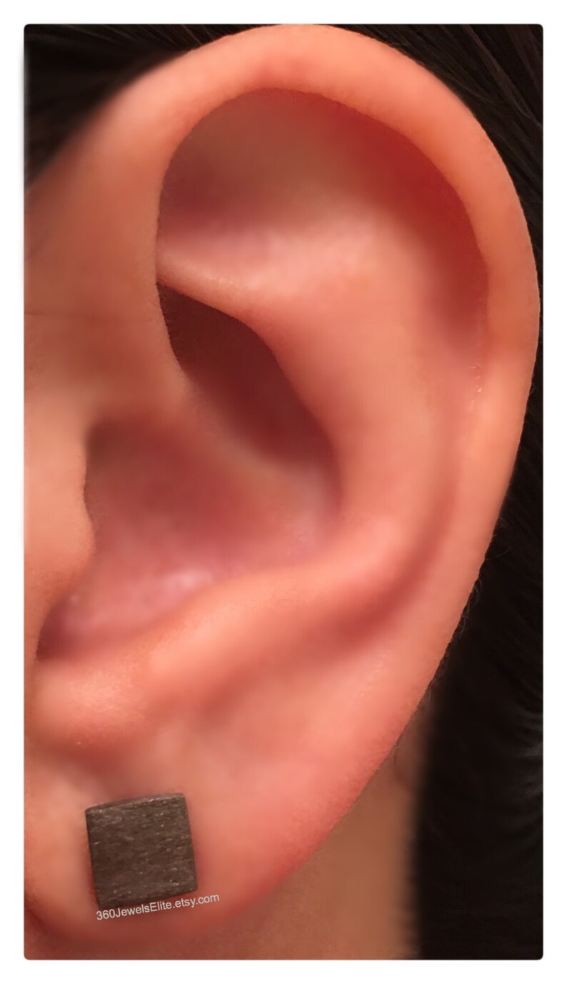 May include: A close-up of a person's ear with a small, square, dark grey earring. The earring is made of wood and has a natural, rustic look. The text "360JewelsElite.etsy.com" is visible below the earring.