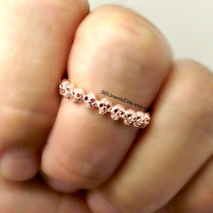 Könnte beinhalten: Roségoldener Totenkopfring mit einer Reihe kleiner Schädel. Der Ring wird an einem Finger getragen.