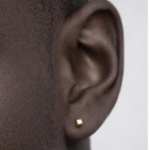 May include: A close-up of a person's ear with a small, square, gold-colored stud earring. The earring is a simple cube shape, and the skin tone is dark. The background is a plain, light color.