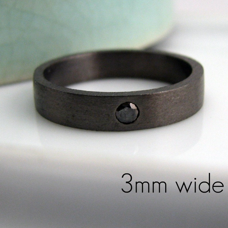 May include: A dark gray ring with a single black gemstone. The ring is 3mm wide and has a brushed metal finish. The ring is a simple, modern design.
