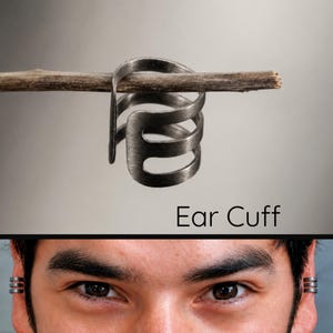 May include: A dark gray metal ear cuff featuring three parallel bands connected by a vertical bar. The text Ear Cuff is printed in black below the jewelry. The ear cuff is shown worn on the upper cartilage of both ears, displaying the triple-band design.