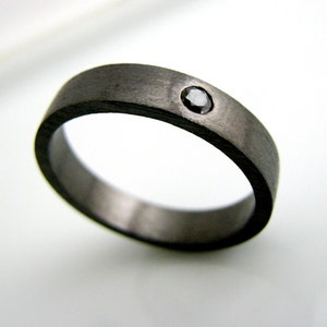 May include: A dark gray ring with a matte finish. The ring features a single, round, black gemstone set into the band. The ring is a simple band design, suitable for everyday wear.