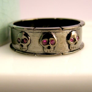 May include: A black metal ring with three skull designs, each with pink gemstone eyes.