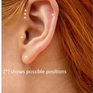 May include: Close-up of a person's ear with multiple white asterisks marking possible piercing locations. The text '[*] shows possible positions' is visible below the ear.