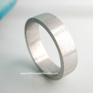 May include: A brushed silver wedding band with a flat, wide profile. The band is smooth and has a matte finish.