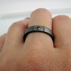 May include: A dark gray ring with a single black gemstone set in the center. The ring has a brushed metal finish and is worn on a finger. The ring is a simple band style.