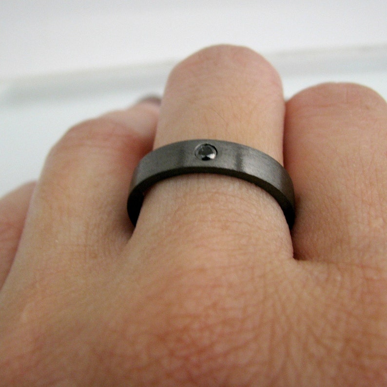 May include: A dark gray ring with a single black gemstone set in the center. The ring has a brushed metal finish and is worn on a finger. The ring is a simple band style.