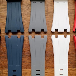 May include: A collection of watch straps in various colors, including navy blue, gray, and white. The straps are made of a flexible material and feature a textured design with multiple holes for adjustment. The straps are displayed on a wooden surface.