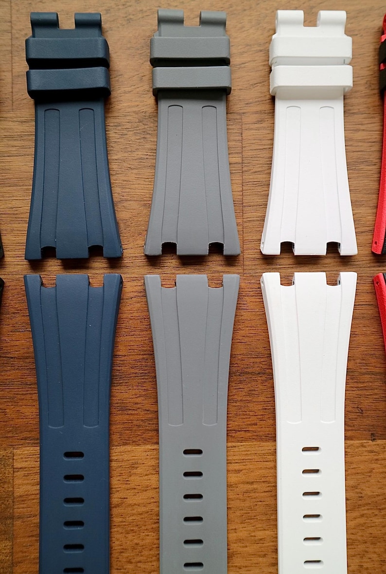 May include: A collection of watch straps in various colors, including navy blue, gray, and white. The straps are made of a flexible material and feature a textured design with multiple holes for adjustment. The straps are displayed on a wooden surface.
