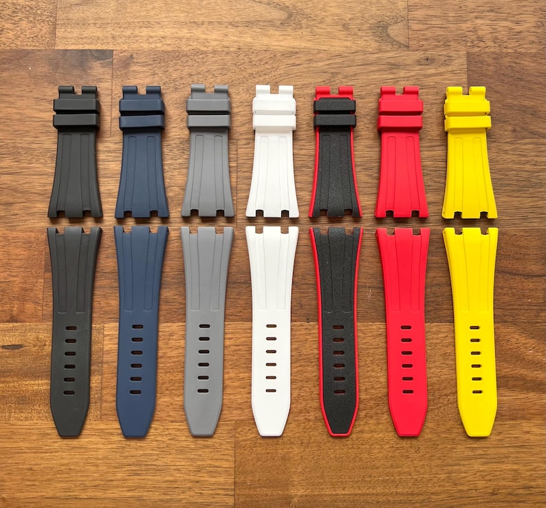 May include: A collection of watch straps in various colors, including black, navy blue, gray, white, black and red, red, and yellow. The straps are made of a flexible material and have a textured design. The straps are displayed on a wooden surface.