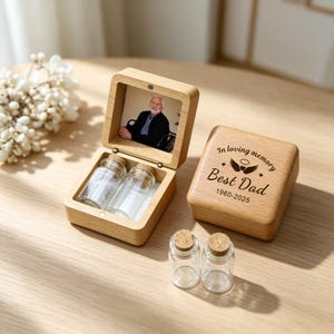 May include: A wooden memorial box with a photo inside, two small glass bottles, and a separate wooden box with the text "In loving memory Best Dad 1960-2025". The open box displays the photo and bottles.