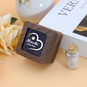 May include: A wooden pet memorial box with a heart-shaped design and the name "Mochi" and dates "2009-2024". A small glass jar with a cork lid and a white book are also in the image. The box is square and made of dark wood.