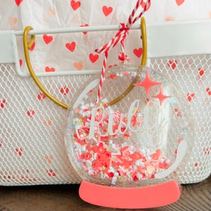 May include: A clear, round ornament with the name "Ruby" inside, filled with red, white, and iridescent confetti. The ornament has a coral-colored base and is suspended by a red and white string. The background includes a white basket and tissue paper with red hearts.