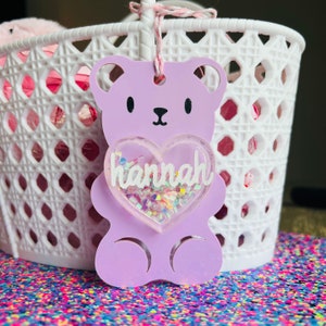 May include: A lavender bear-shaped tag with a heart-shaped center containing the name "hannah" in white script. The heart is filled with iridescent confetti. The tag is hanging in front of a white basket.