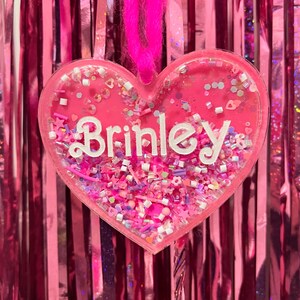 May include: A pink heart-shaped ornament with the name "Brinley" in white script. The ornament is filled with pink and iridescent confetti and has a pink ribbon for hanging. The background is a pink, shiny, vertical curtain.