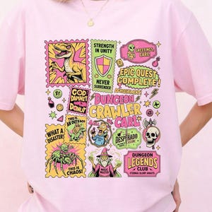 May include: A light pink t-shirt featuring a colorful graphic design with various illustrations and text elements. The design includes images of a dinosaur, shield, skull, and other fantasy-themed elements. Text includes "Dungeon Crawler Care" and "Epic Quest Complete!"