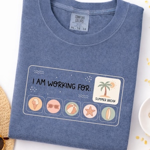 May include: A blue Comfort Colors t-shirt with the text "I AM WORKING FOR: SUMMER BREAK" and summer-themed icons. The shirt is folded and lies on a white surface, with a straw hat and sunglasses in the corner.