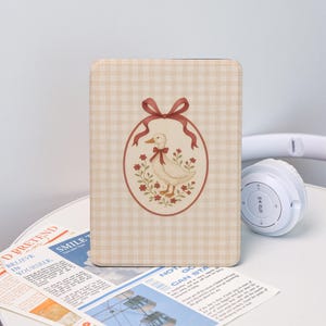 Könnte beinhalten: Eine rechteckige Hülle mit beigem Vichy-Muster. Die Hülle zeigt eine ovale Illustration einer Ente mit Schleife, umgeben von Blumen und einem roten Band. Ein weißer Kopfhörer liegt in der Nähe.