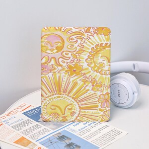 May include: A yellow and white patterned tablet case featuring a sun and floral design. The case is rectangular and has a soft, matte finish. A pair of white headphones is visible in the background.