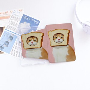 Könnte beinhalten: Eine rechteckige Schutzhülle für ein elektronisches Gerät mit einer Grafik einer orange-weißen Katze, deren Kopf durch eine Scheibe Toastbrot schaut. Der Hintergrund der Hülle ist in einem einfarbigen, gedeckten Rosaton gehalten.