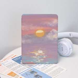 May include: A rectangular tablet case featuring a sunset scene with a yellow sun, clouds, and water reflections in shades of pink, purple, and blue. White headphones are visible on the right. The case is on a white surface with newspaper clippings.