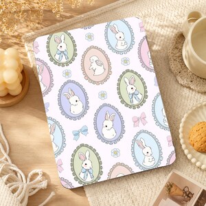 May include: A rectangular tablet case featuring a repeating pattern of white rabbits inside oval frames of various pastel colors, including green, pink, blue, and purple, interspersed with small blue flowers and light pink and blue bows.