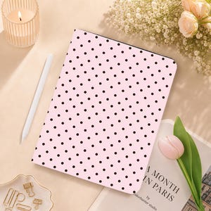 May include: A rectangular tablet case featuring a light pink background with a repeating pattern of small black polka dots. The case is positioned alongside a white stylus, a candle in a ribbed glass holder, a book, and a single pink tulip.