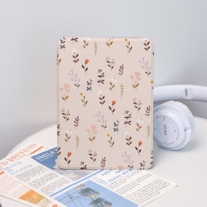 May include: A rectangular tablet case with a beige background and a repeating floral pattern in shades of brown, orange, and white. The case is on a white surface next to a pair of white headphones and a newspaper.