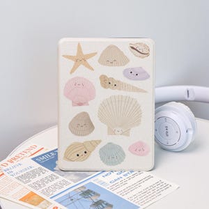May include: A white tablet case featuring a collection of smiling seashell illustrations in various colors, including pink, beige, and blue. A starfish is also included in the design. A pair of white headphones rests on the table.