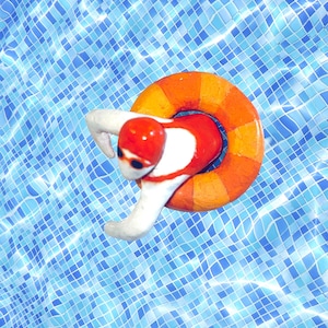 Peut inclure: Une figurine rouge et blanche d'une personne portant un bonnet de bain et tenant un anneau gonflable orange et jaune. La figurine flotte dans une piscine aux carreaux bleus.