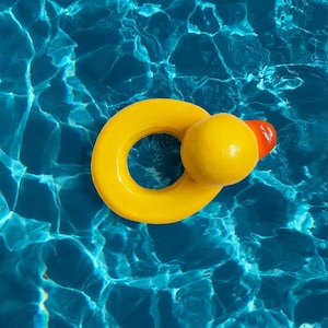Peut inclure: Un anneau gonflable jaune en forme de canard avec un bec orange flotte dans une piscine d'eau bleue. Le flotteur de piscine est de couleur jaune vif et l'eau a une surface ondulée.