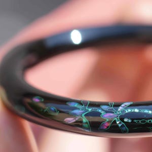 May include: Black bangle bracelet with a dragonfly design made from iridescent abalone shell inlay.