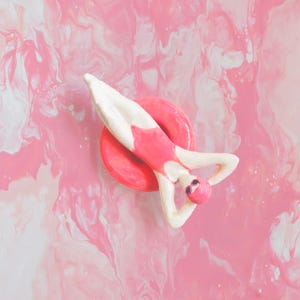 Könnte beinhalten: Eine skurrile Keramikfigur einer Schwimmerin in einem rosa Badeanzug und einer Badekappe, die auf einem rosa Schwimmring ruht. Die Figur ist in Rosa- und Weißtönen bemalt, mit Sonnenbrille. Der Hintergrund ist ein rosa-weißes Marmormuster.