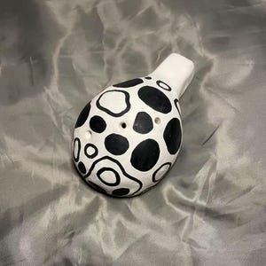 Può includere: Ocarina in ceramica bianca con un motivo a macchie nere. L'ocarina ha un corpo arrotondato con diversi fori per le dita e un bocchino. Le macchie nere variano per dimensioni e forma, creando un design unico. Lo strumento è progettato per essere tenuto e suonato.
