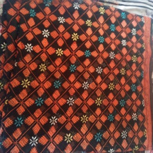 Puede incluir: Un textil negro con un intrincado patrón de motivos florales y de hojas en naranja, blanco, amarillo y turquesa. El diseño se repite en toda la tela, creando un efecto geométrico. Los bordes están adornados con un borde naranja.