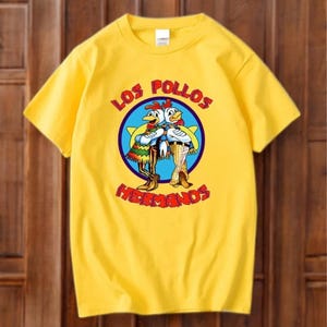 May include: Yellow t-shirt with a cartoon image of two chickens in a blue circle, with the words "LOS POLLOS HERMANOS". The chickens are wearing outfits and standing together. The shirt is made of a soft material.