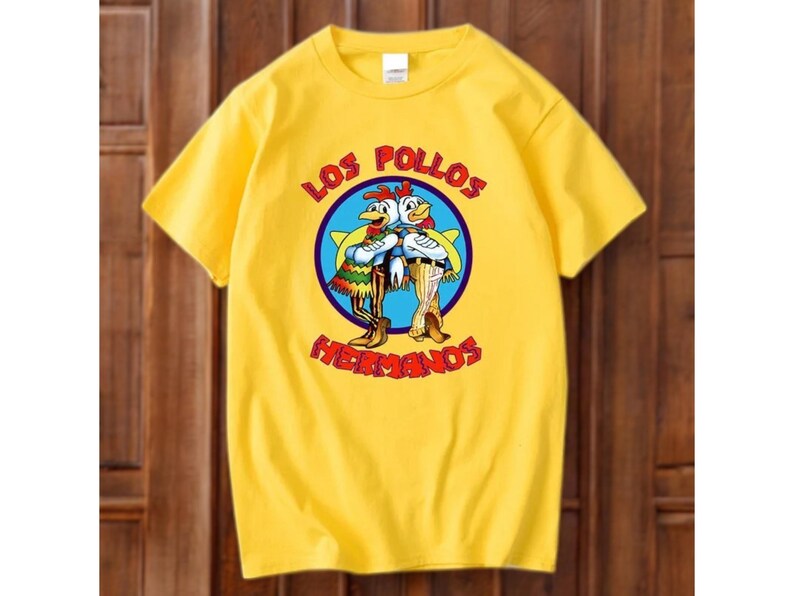 May include: Yellow t-shirt with a cartoon image of two chickens in a blue circle, with the words "LOS POLLOS HERMANOS". The chickens are wearing outfits and standing together. The shirt is made of a soft material.