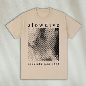 K&ouml;nnte beinhalten: Ein beiges T-Shirt mit einem schwarz-wei&szlig;en Grafikdruck eines Gesichts. Der Text "slowdive" und "souvlaki tour 1994" ist auf dem Shirt aufgedruckt.