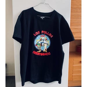 May include: Black t-shirt with a cartoon graphic of two chickens and the text "LOS POLLOS HERMANOS". The design is in red, white, and blue. The t-shirt is hanging on a metal hanger.