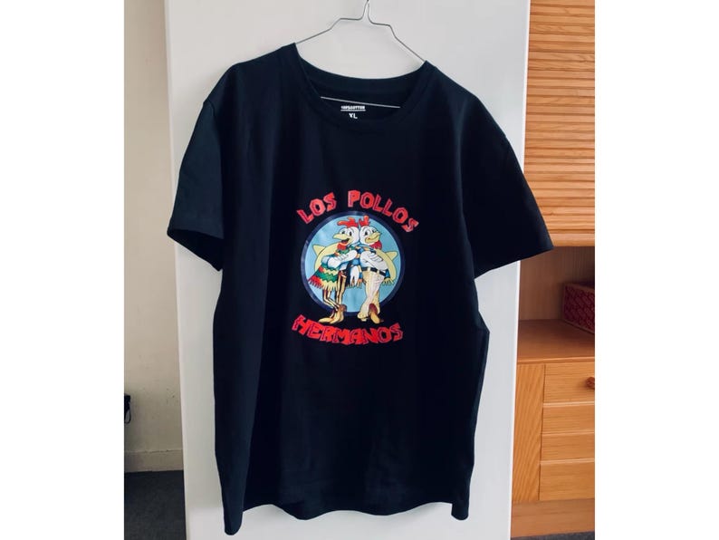 May include: Black t-shirt with a cartoon graphic of two chickens and the text "LOS POLLOS HERMANOS". The design is in red, white, and blue. The t-shirt is hanging on a metal hanger.