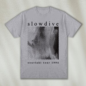K&ouml;nnte beinhalten: Graues T-Shirt mit schwarzem Text, der "slowdive" und "souvlaki tour 1994" lautet. Das Shirt zeigt ein Schwarzwei&szlig;-Halbtonbild.